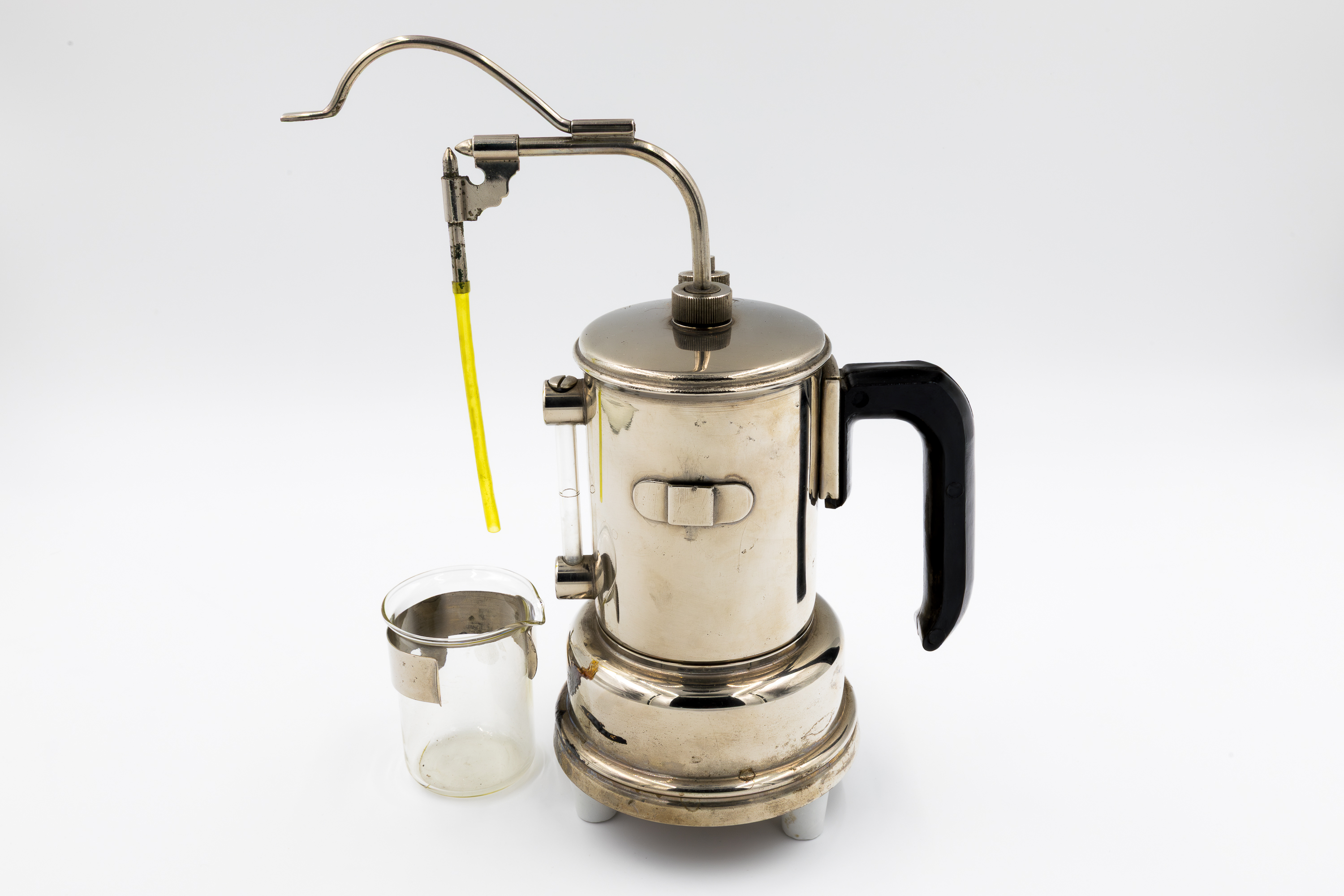 Na zdjęciu widać metalowy, elektryczny sterylizator medyczny z połyskującą, chromowaną powierzchnią. Urządzenie ma cylindryczny kształt, czarny uchwyt z boku i wylot pary zakończony metalową rurką z żółtym przewodem. Obok sterylizatora stoi przezroczysta szklana zlewka laboratoryjna. Całość stoi na białym tle i przypomina niewielki czajnik elektryczny, używany dawniej do sterylizacji narzędzi medycznych w placówkach ochrony zdrowia.